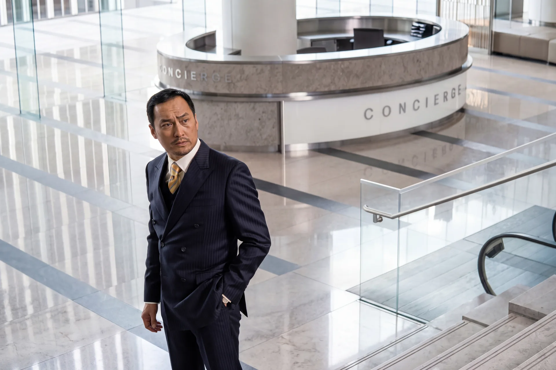 A man in a pinstripe suit stands in a hotel lobby beside a concierge desk.